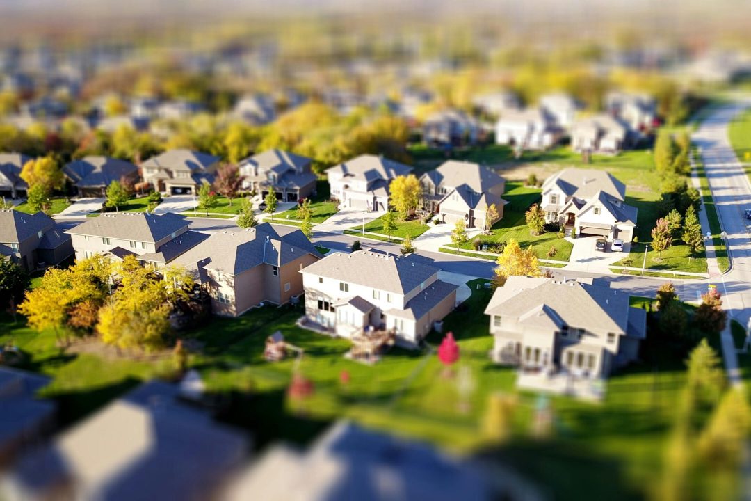 Tilt-shift aerial photo of a suburban neighborhood with green lawns and roads.
