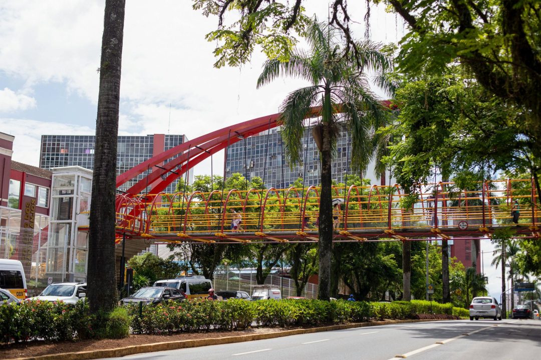 Colorful footbridge over city street in Joinville, Brazil, surrounded by lush greenery and modern buildings.