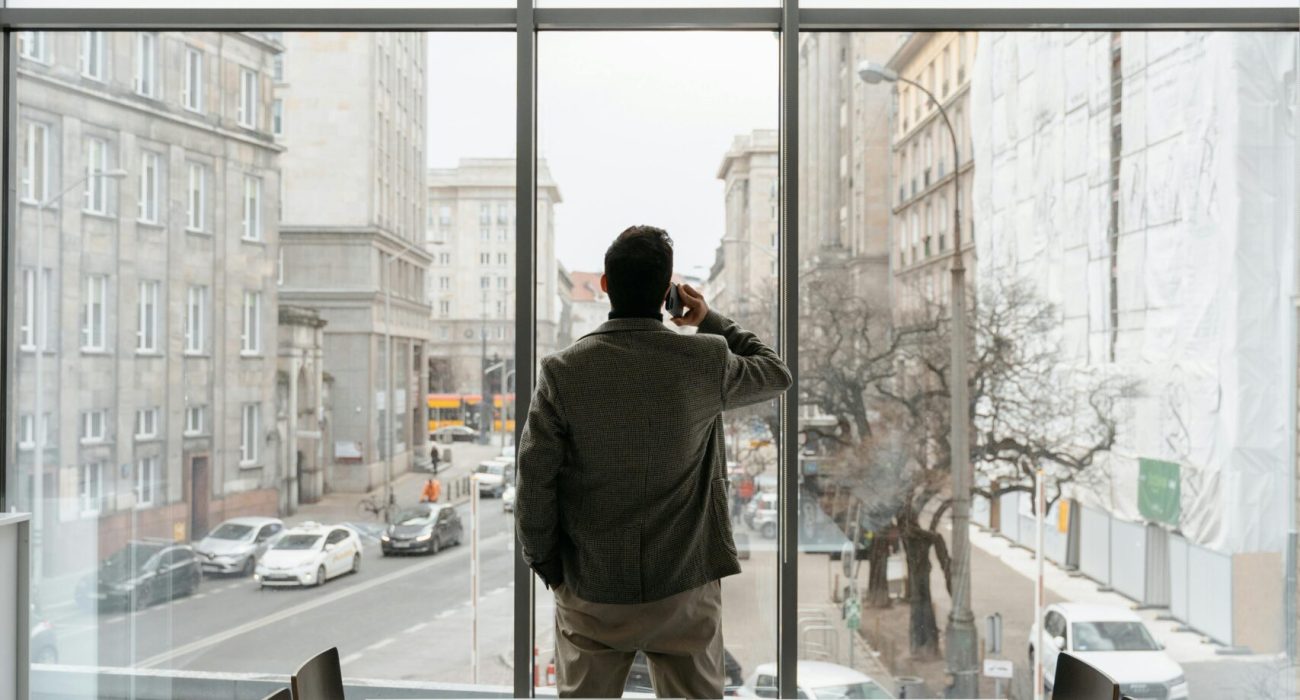 Businessman standing in a modern office, talking on the phone while looking out the window onto a city street.